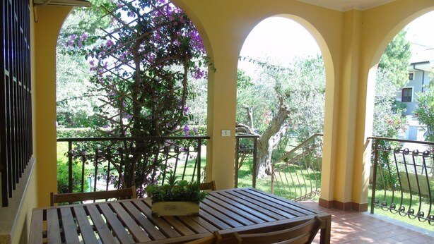 A covered patio with a wooden table and chairs, surrounded by yellow walls and a view of greenery and trees.