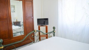 A bedroom with a wooden wardrobe, a small wooden table with a lamp, and a television on a stand.