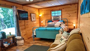 Acogedora habitación tipo cabaña con cama, sofá, televisor y ventana con vistas a árboles.