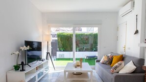 Een moderne woonkamer met een grijze bank, een witte salontafel en een flatscreen televisie op een witte dressoir. Een grote schuifpui leidt naar een groene tuin.