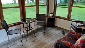 A room with large windows, a glass table, two chairs, a small side table with a lantern, and a colorful sofa.