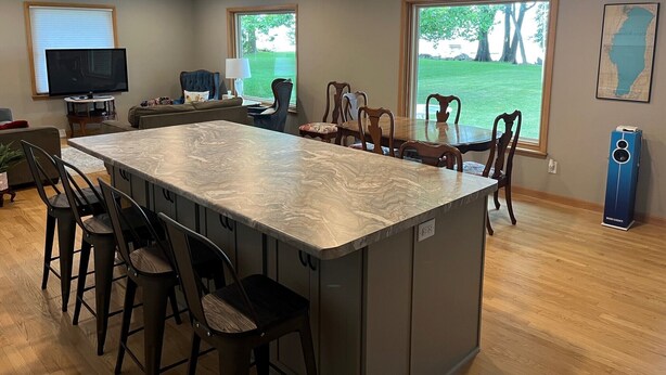 A modern kitchen with a marble island, bar stools, and a large window offering a view of the outdoors.