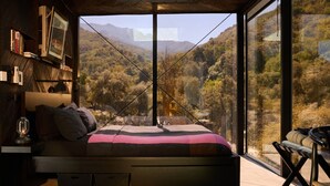 A bedroom with a bed, bedside tables, a lamp, and a view of a forested landscape through large windows. - Paradise Ranch Inn- Infinite House Hot Tub,Sauna. (Three Rivers)