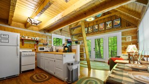A rustic cabin interior with wooden beams, a kitchen with a stove, refrigerator, and shelves, and a dining area with a table set for two.