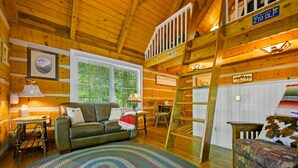A cozy log cabin interior with a loft bedroom, a dining area, and a living room with a sofa and a coffee table.
