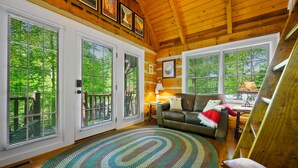 A cozy cabin interior with a wooden ceiling, large windows with blinds, a leather sofa, a patterned rug, and a small table with a lamp.