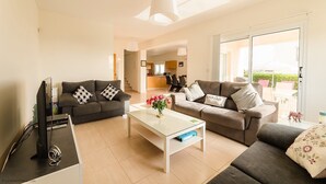 A modern living room with a sofa, a coffee table, and a TV. There is a kitchen in the background and a sliding glass door leading to an outdoor area. - 3 Bedroom Villa with Private Pool and Free Car (Paphos)
