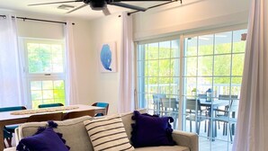 A living room with a beige sofa, two purple pillows, a blue patterned rug, a wooden coffee table with books, and a ceiling fan. - Intimate remodeled villa with private beach and pool. (Providenciales)