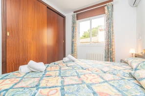 Una habitación de hotel con un armario de madera, una cama con una colcha estampada y colorida, una ventana con cortinas y una lámpara de noche.