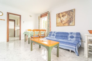 Una sala de estar con un sofá, una mesa de madera y dos sillas. Hay un cuadro en la pared y una ventana con cortinas.
