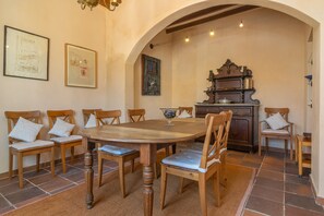 A dining area with a round wooden table surrounded by six wooden chairs with cushioned seats, a wooden cabinet with a decorative top, and a tiled floor.
