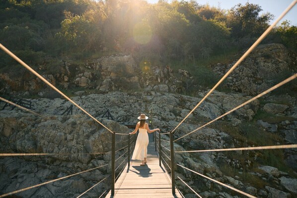 A wooden suspension bridge with metal railings, leading to a rocky landscape with trees.