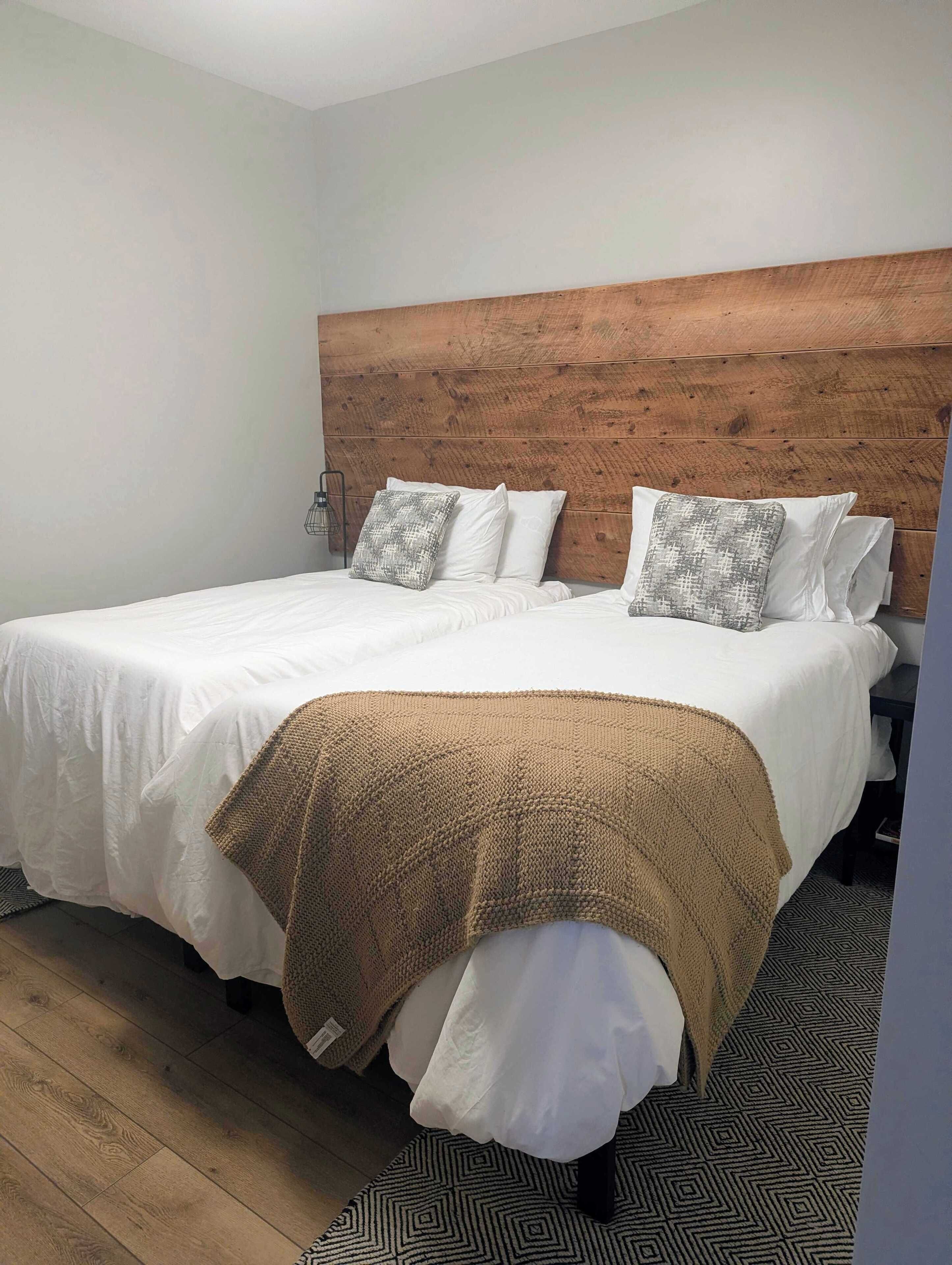 Ein ordentlich bezogenes Bett mit weißen Leinen, zwei grauen Kissen, einem Holzkopfstück und einer braunen Strickdecke.