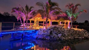 A nighttime resort with a pool, palm trees, and a thatched-roof structure. - VENUE EVENTS Cbc Farms 100 person (Okeechobee)
