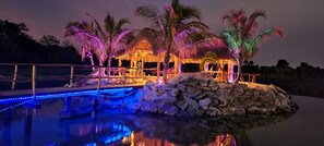Un complexe hôtelier ouvert la nuit, doté d’une piscine, de palmiers et d’une structure à toit de chaume.