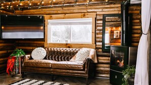 L’intérieur chaleureux d’un chalet avec un canapé en cuir, un téléviseur à écran plat, une cheminée et une fenêtre donnant sur un paysage enneigé.