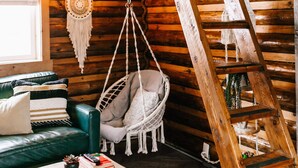 L’intérieur d’un chalet rustique, avec un lit en mezzanine, un fauteuil suspendu, une échelle en bois et un coin salon confortable agrémenté de coussins.