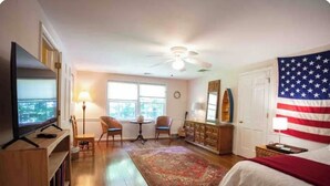A hotel room with a television, a ceiling fan, a large American flag, a bed, a wooden dresser, and a window with a view. - Fox Knoll 1-Studio-Sleeps 2-Beautiful Setting! Indy 500! Grand Park, Concerts! (Indianapolis)