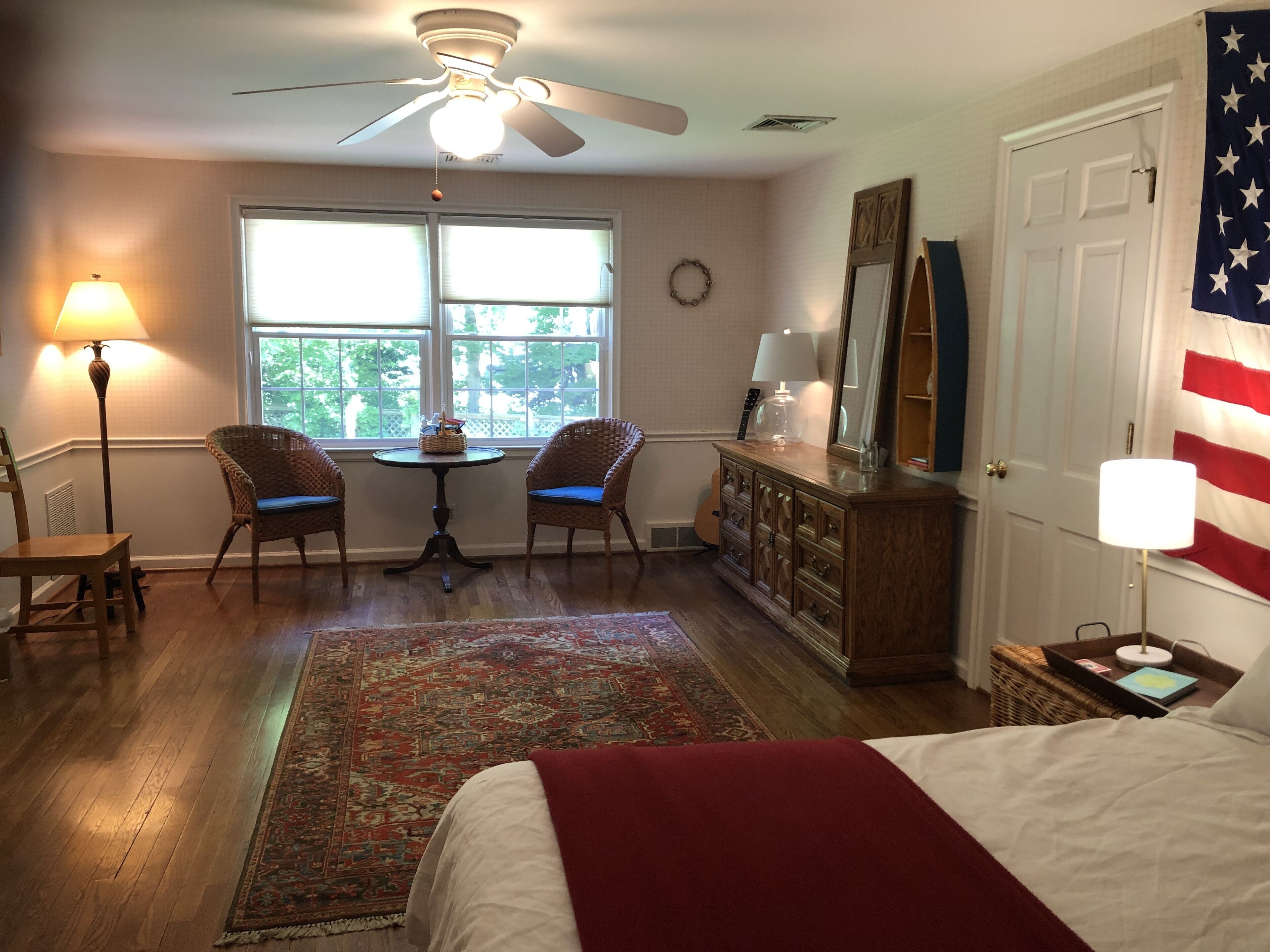 A bedroom with a bed, two chairs, a table, a dresser, an American flag, and a ceiling fan.