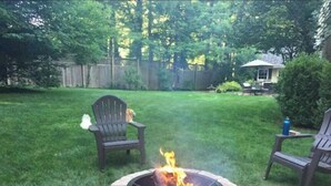 A backyard with a stone fire pit surrounded by grass, two Adirondack chairs, and a wooden fence in the background. - Fox Knoll 1-Studio-Sleeps 2-Beautiful Setting! Indy 500! Grand Park, Concerts! (Indianapolis)