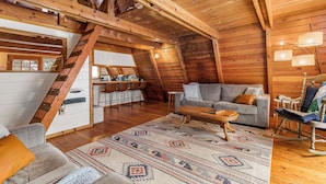 A cozy loft-style room with a wooden ceiling, a staircase, a dining area, and a living space with a sofa and coffee table.