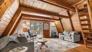 A cozy loft bedroom with a sloped ceiling, wooden beams, a sofa, a chair, a small table, and a ladder.