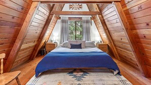 A cozy attic bedroom with a wooden ceiling, a large bed with a blue bedspread, two nightstands, and a window with curtains.