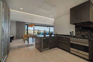 Une cuisine moderne dotée d’un grand îlot central, de meubles de rangement aux couleurs sombres et offrant une vue sur l’océan.