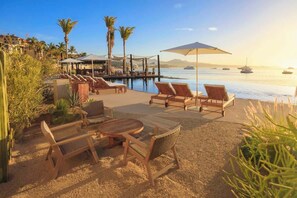 Un salon de plage doté de chaises longues et d’une table, offrant une vue sur une mer calme où voguent des bateaux.