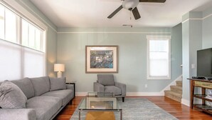 A living room with a gray sofa, a glass coffee table, a gray armchair, a framed picture on the wall, and a staircase in the background. - The Nest - The Perfect Asheville Getaway; Pet Friendly!, 10 Mins to Downtown (Asheville)