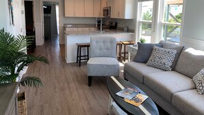 A modern kitchen with a central island, a dining area with a grey upholstered chair, a sofa with patterned cushions, and a ceiling fan. - Newly renovated 3BR/3BA w king bed, linens, towels & views! 🌞 (Surf City)
