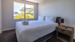 Ein Hotelzimmer mit großem Fenster, einem Bett mit weißen Leinentüchern, einem dunklen Kopfteil und einem kleinen Nachttisch mit Lampe.