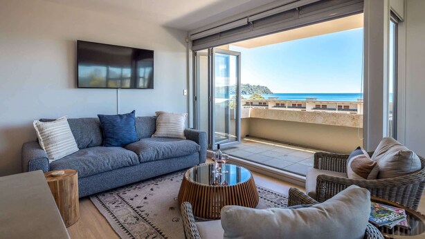 Ein Wohnzimmer mit einer Couch, einem Couchtisch und Korbstühlen. Ein großes Fenster bietet Blick auf das Meer.
