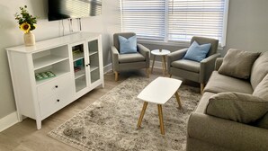 Un salon avec un meuble blanc, deux fauteuils gris, une table basse blanche et une grande fenêtre avec des stores.
