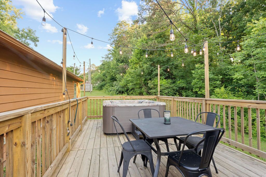 Une terrasse en bois avec un jacuzzi, une table et des chaises, entourée d’arbres et illuminée de guirlandes lumineuses.