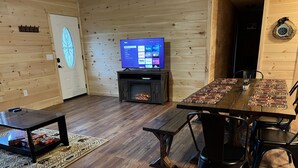 Un saló d'estar acollidor amb interior de fusta, una llar de foc, un televisor i una zona de dinar amb taula i cadires.