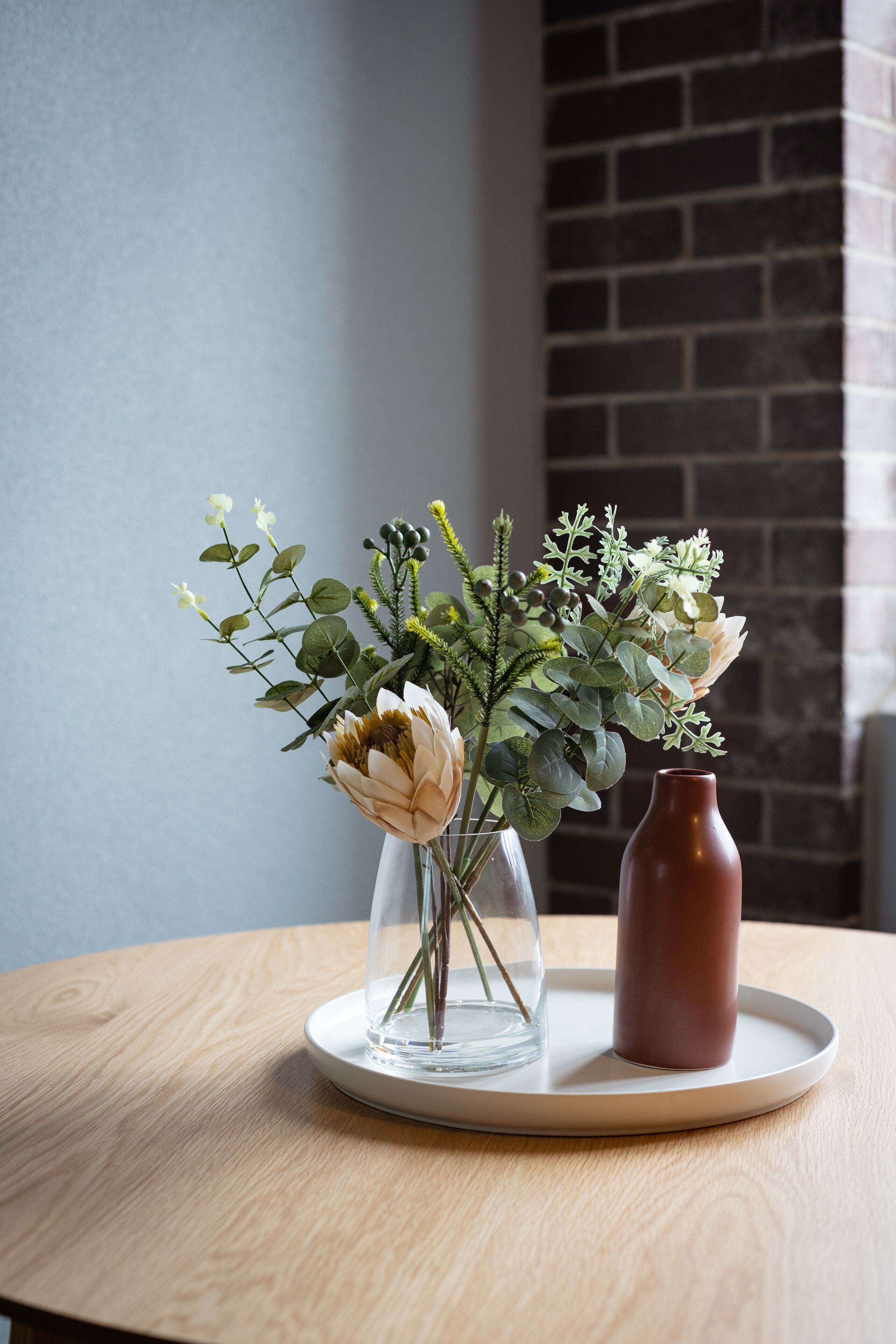 A round wooden table with two vases containing flowers and greenery.