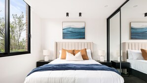 Une chambre d’hôtel moderne avec un grand lit, deux tables de chevet avec des lampes, une grande fenêtre donnant sur les arbres et une œuvre d’art accrochée au mur.