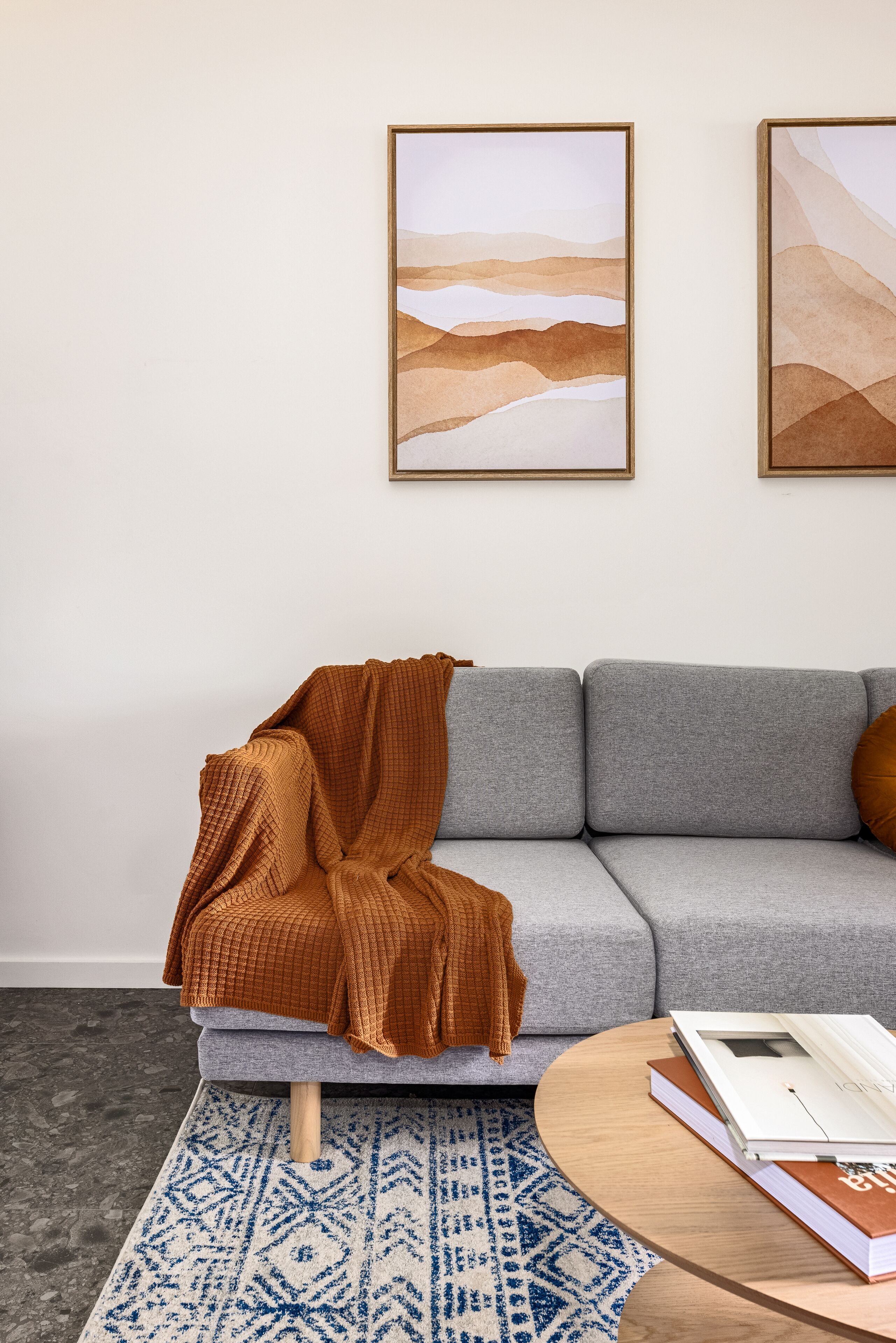 Un salon moderne avec un canapé gris, un plaid marron, une table basse ronde en bois sur laquelle sont posés des livres, et un tapis à motifs.