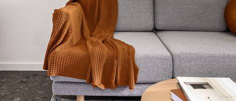 Una sala de estar moderna con un sofá gris, una manta marrón, una mesa de centro redonda de madera con libros y una alfombra estampada.