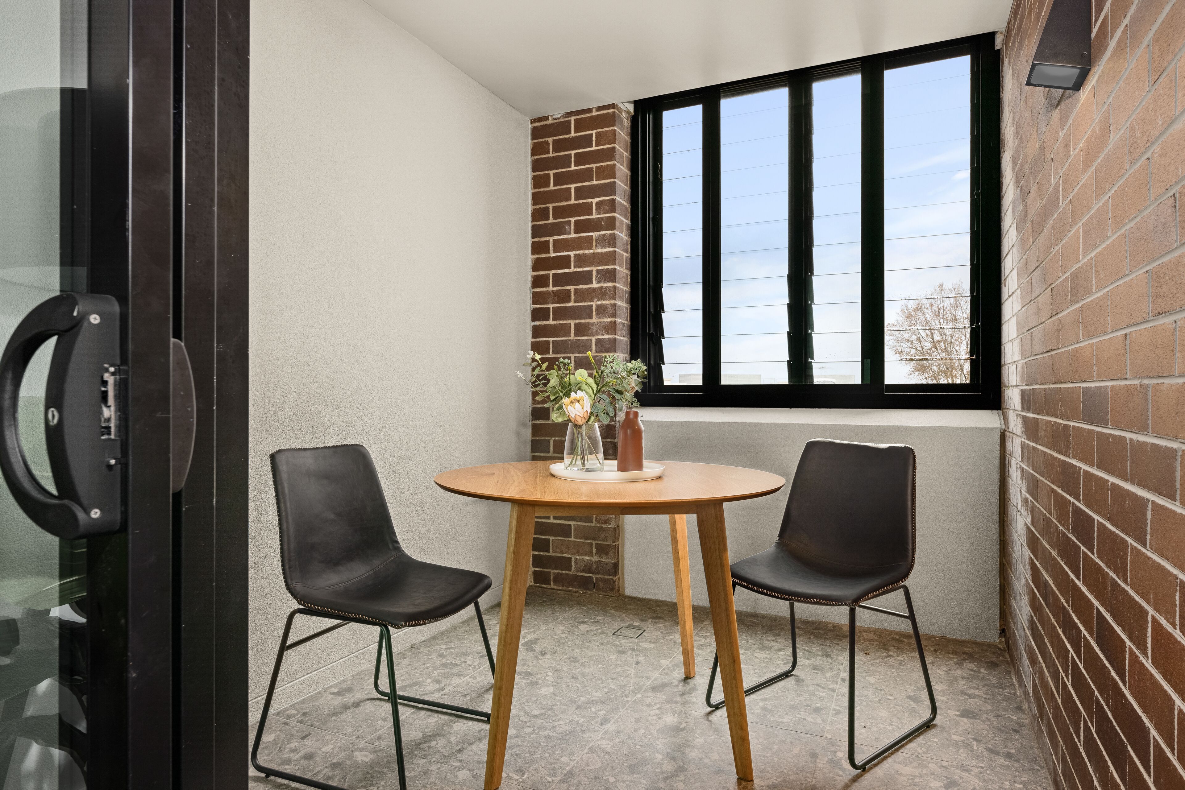 A corner room with a brick wall, a large window, a round wooden table with two black chairs, and a vase with flowers.
