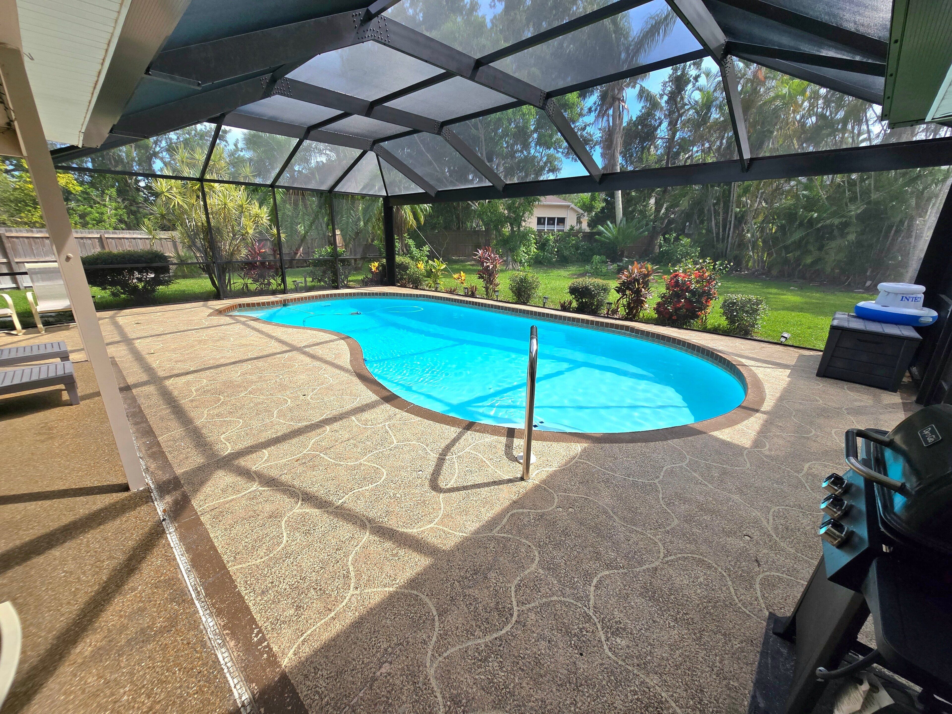 Piscina exterior coberta com um lago azul cristalino, rodeada por um jardim ajardinado e vista para uma área residencial.
