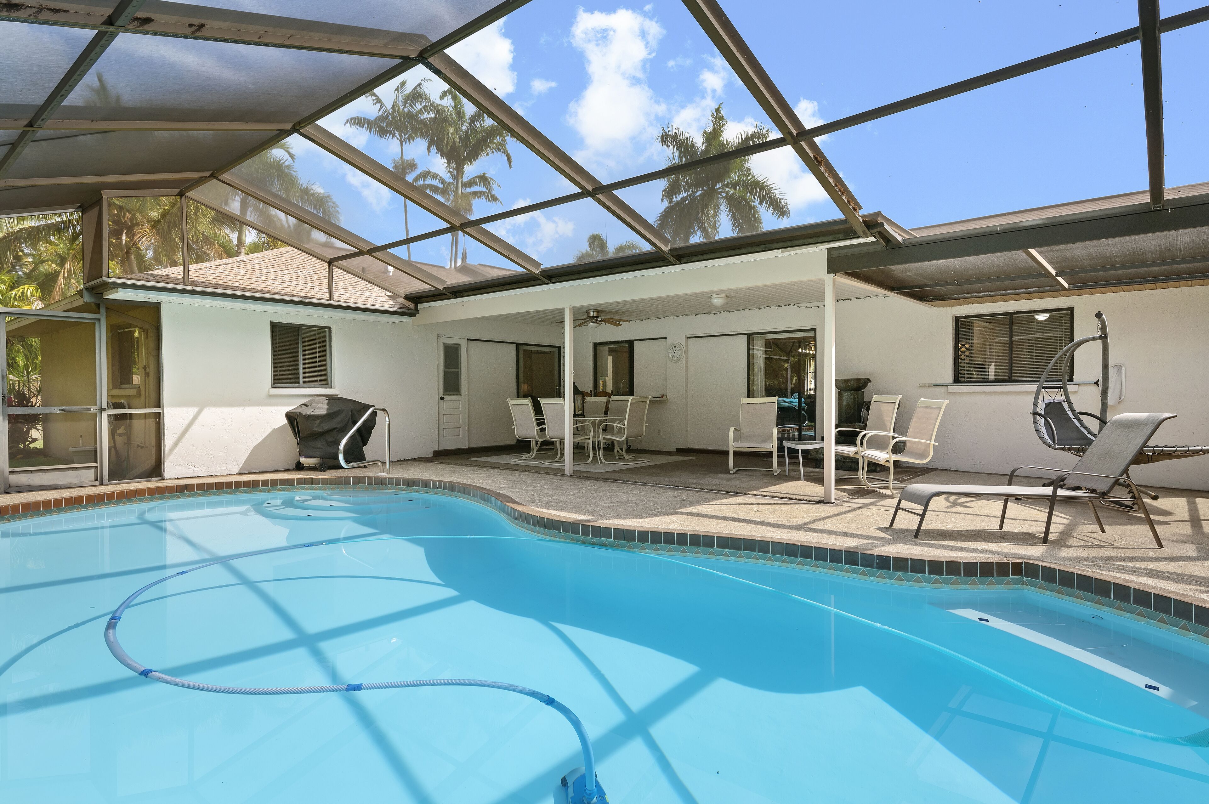 A swimming pool with a glass ceiling, surrounded by white chairs and a table, with palm trees visible outside.