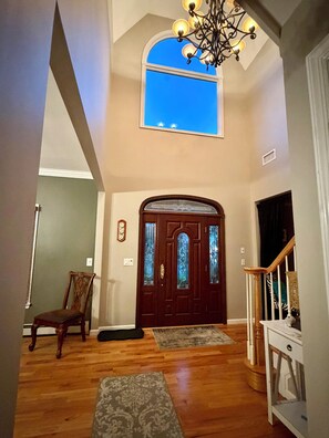 A spacious foyer with a wooden floor, a dark wooden door with glass panels, a chandelier, and a view of the sky through a window.