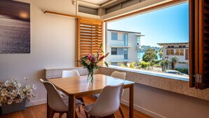 A dining area with a wooden table and white chairs, a window with wooden blinds, and a view of buildings and trees.