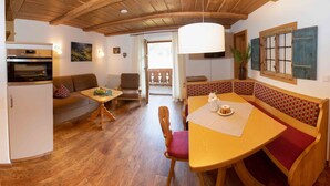 A cozy living room with a wooden ceiling, a dining table with a bench, a sofa, and a kitchen area with a refrigerator and oven. - 4 Fewo , Balkon, Kochnische, 1 Schlaf- und 1 Wohn-/schlafz. max 4 Pers, Wlan (Ruhpolding)