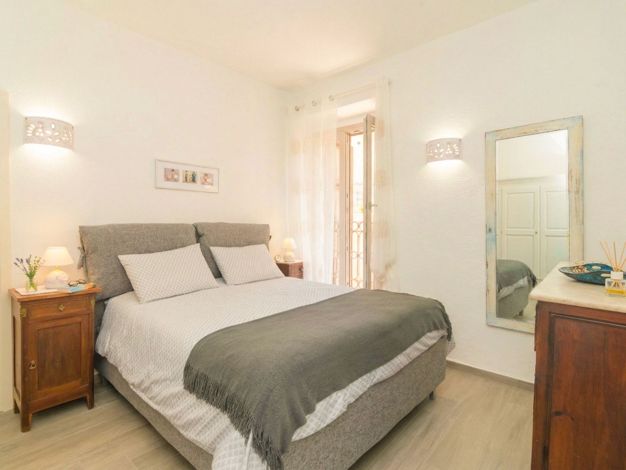 A bedroom with a bed, bedside tables, a mirror, and a door leading to another room.