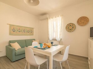 Un coin repas avec une table blanche, deux chaises blanches, un canapé vert et une fenêtre ornée de rideaux légers.