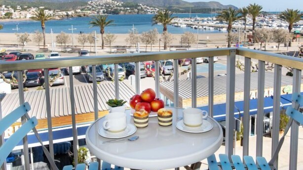 A balcony with a table set for two, overlooking a marina with boats and a coastal town.