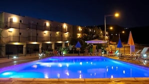 Una piscina con borde iluminado en azul, rodeada de sillones de descanso y sombrillas, con un edificio al fondo.